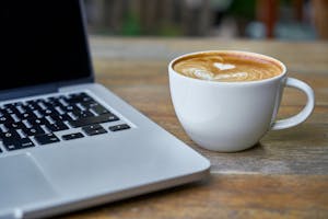 Close-up of a latte coffee with heart art next to a laptop on a rustic wooden table.